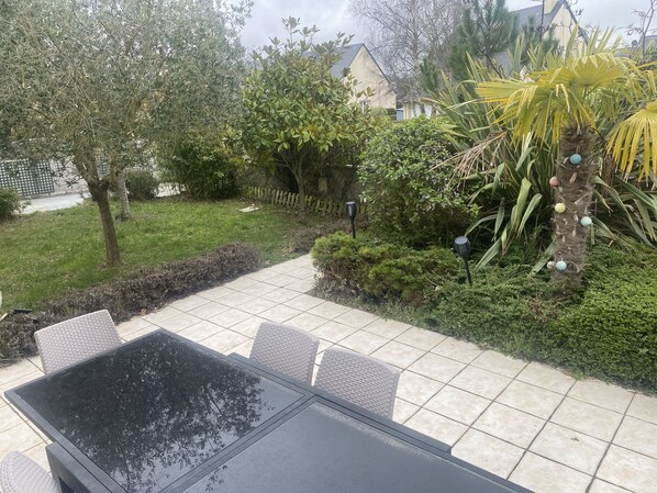 A patio with a glass-top table and wicker chairs, surrounded by a garden with various plants and a palm tree. - Large, quiet house 5 minutes' walk from the beach (Cabourg)