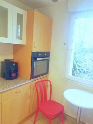 A small kitchen with wooden cabinets, a coffee maker, a red chair, and a window with a view of trees. - Large, quiet house 5 minutes' walk from the beach (Cabourg)