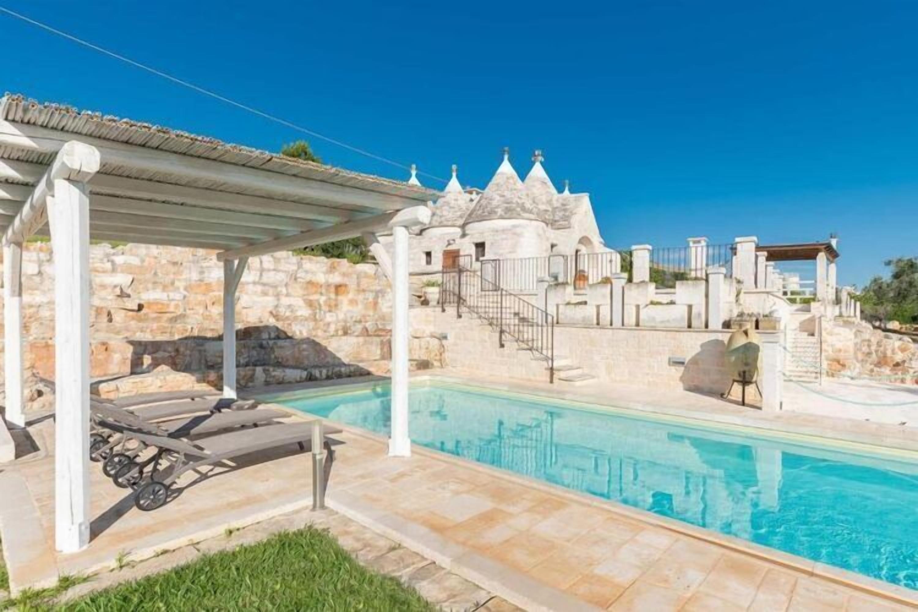 A swimming pool with lounge chairs and a pergola, surrounded by stone walls and a staircase leading to a building with a unique architectural style.