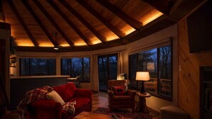 Una sala de estar acogedora con un sofá rojo, una mesa de centro de madera y un sillón con una lámpara. Amplias ventanas ofrecen vistas al exterior.