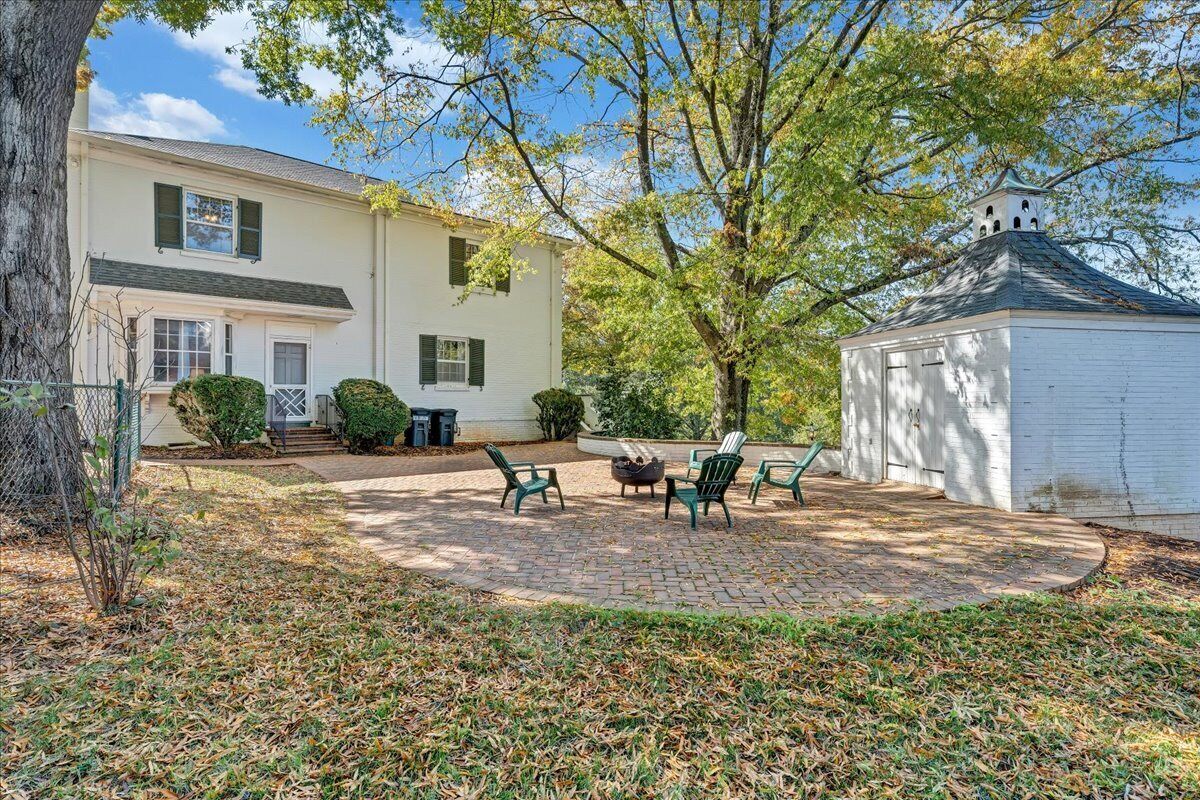 A white building with a chimney, a covered patio with chairs and a fire pit, and a tree in the background.