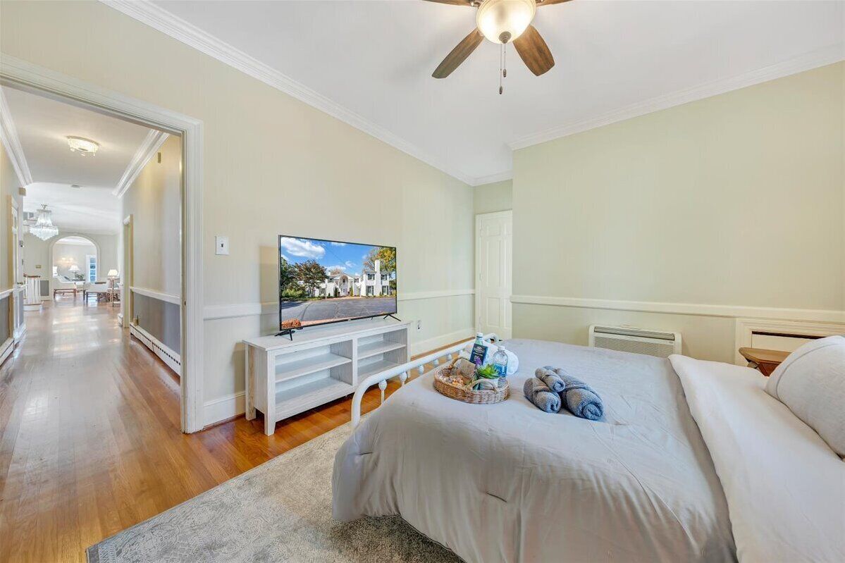 A bedroom with a bed, a TV on a stand, a ceiling fan, and a view of a street.