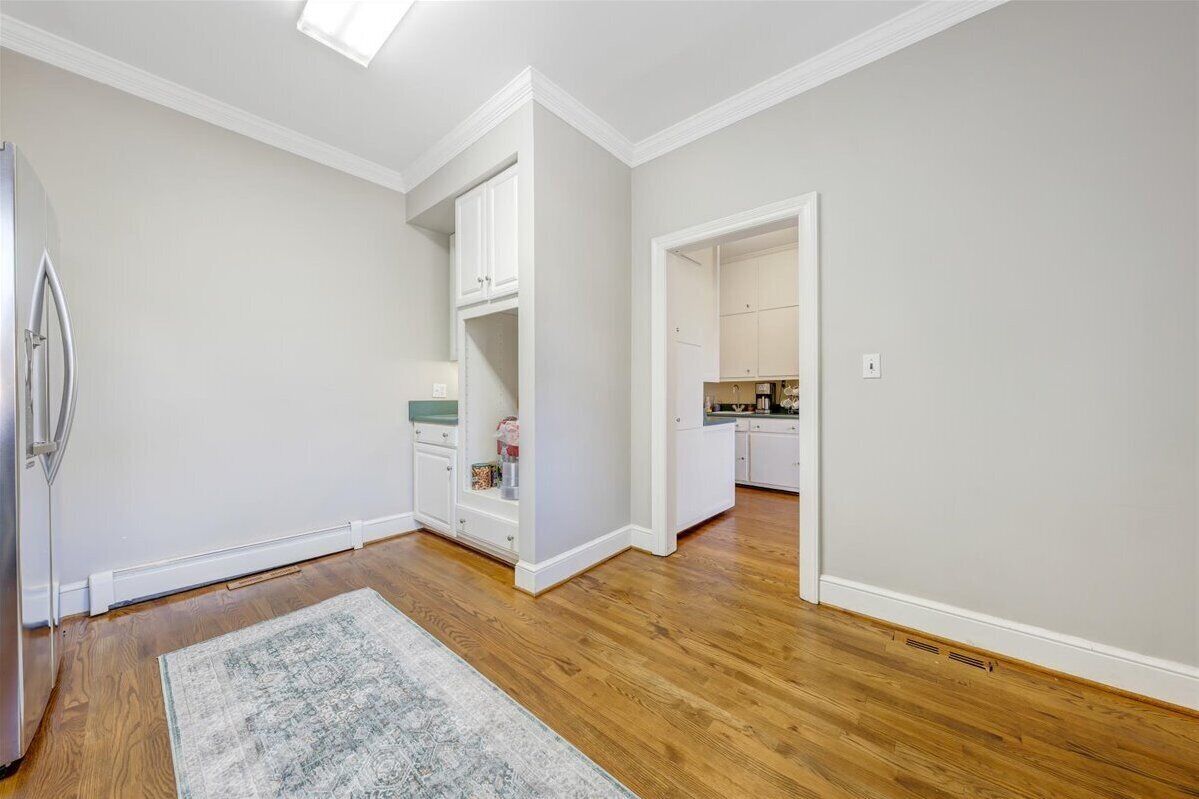 A spacious room with hardwood flooring, a built-in white cabinet, and a kitchen area visible through an open doorway.
