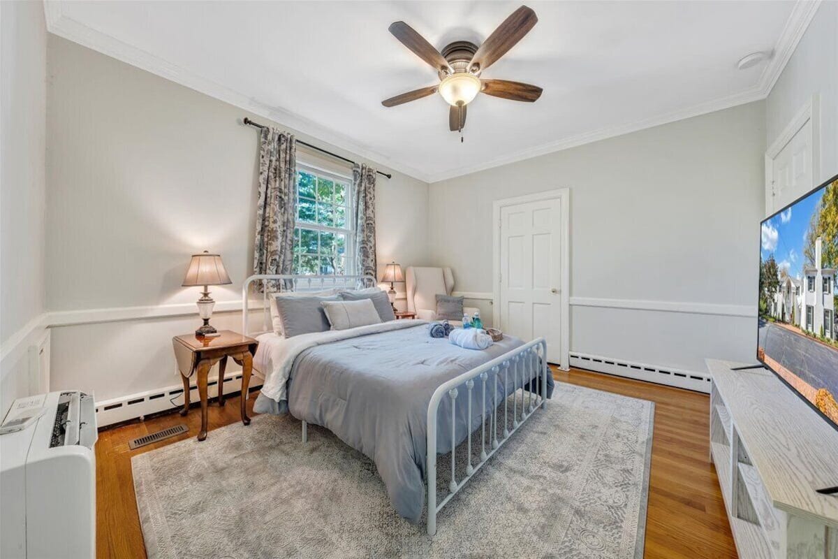 A bedroom with a bed, bedside lamps, a ceiling fan, a TV, and a window with curtains.