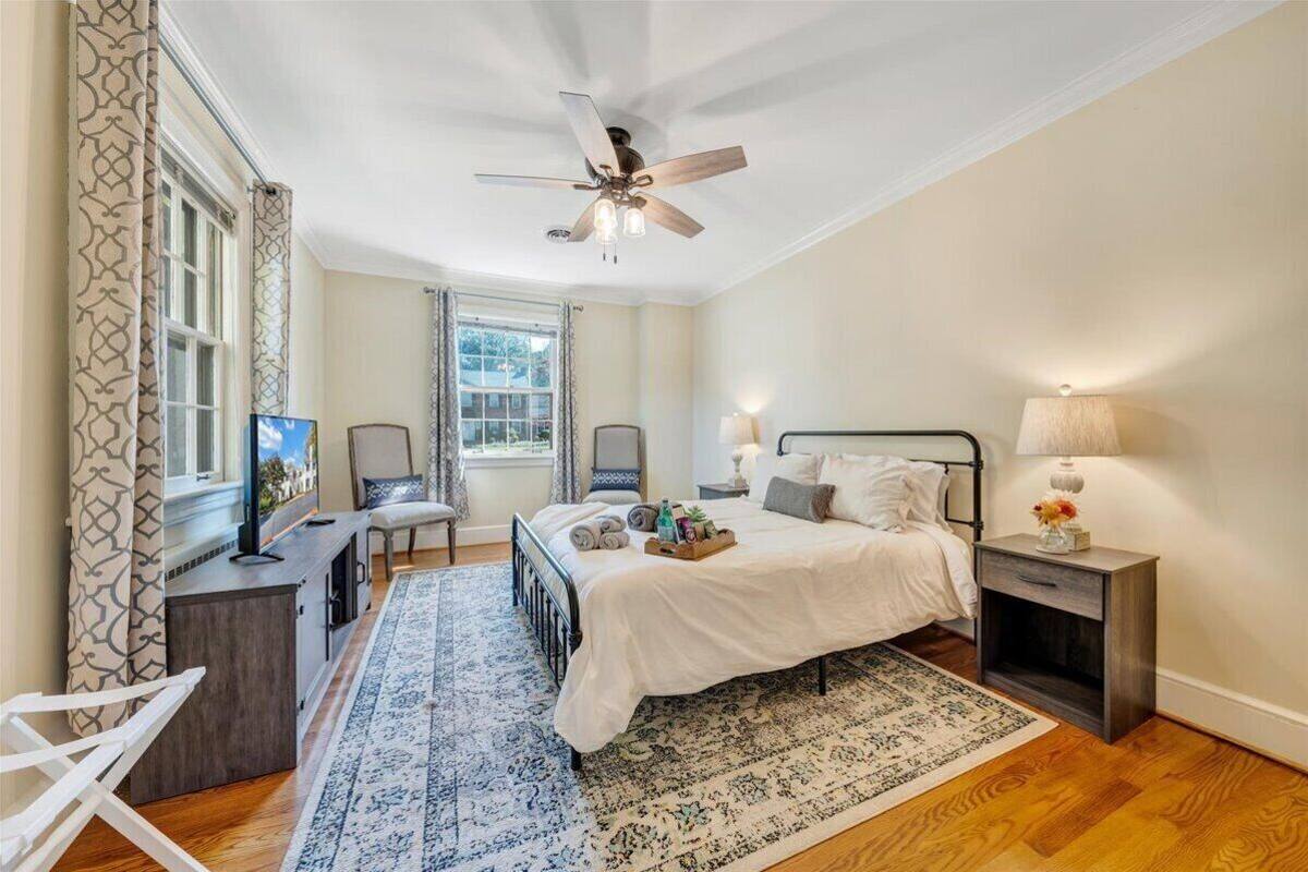 A bedroom with a bed, bedside tables, a desk, a chair, a ceiling fan, and a window with curtains.