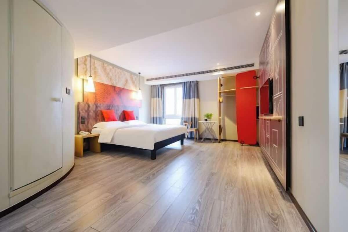 A modern hotel room with a large bed, a wardrobe, a small table with a lamp, and a view of the hallway.