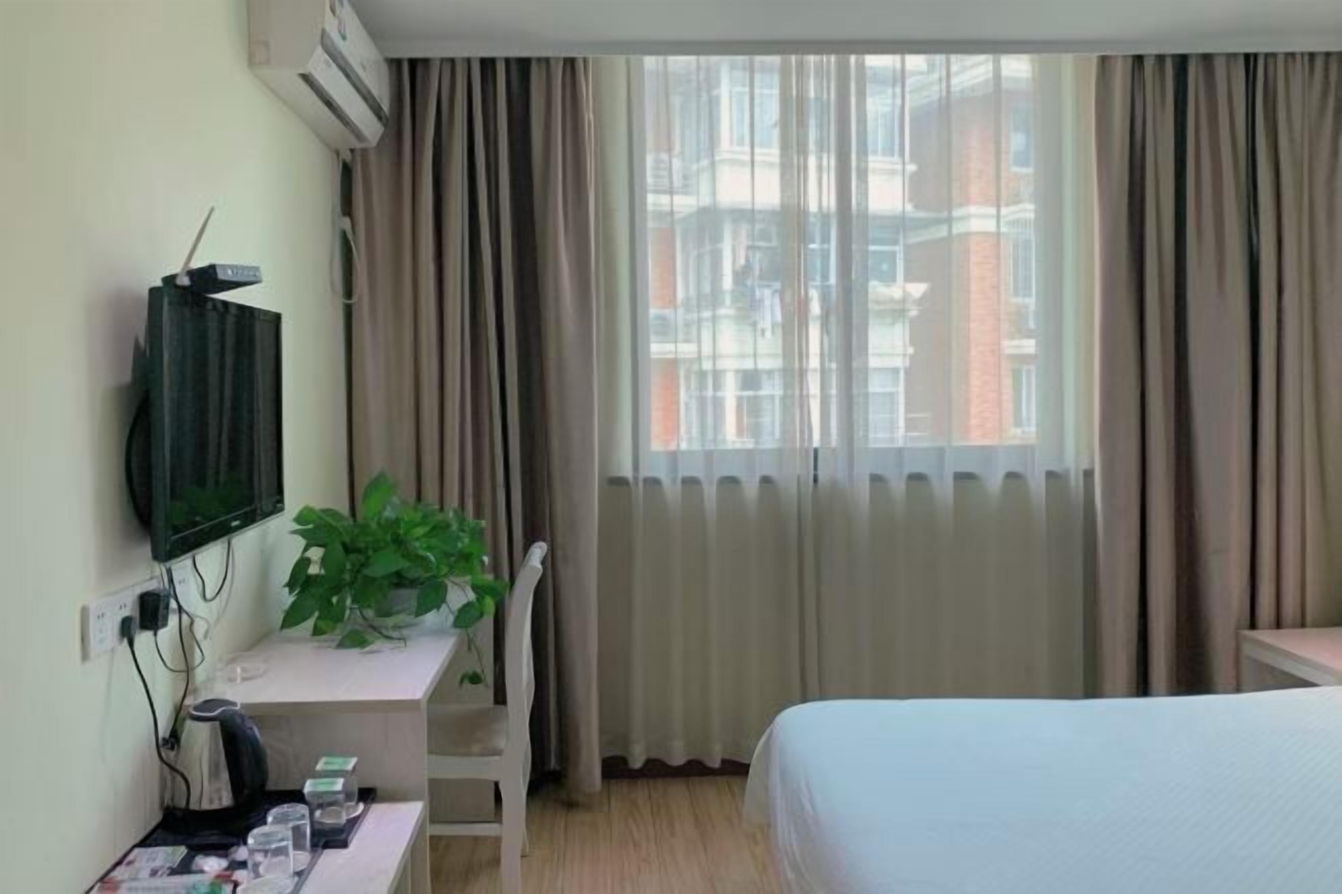 A hotel room with a bed, a desk with a plant, a TV, and a window with curtains.
