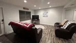 A living room with a dark wood floor, a red and white blanket on a recliner, a black leather sofa with a blanket, a flat-screen TV, and a small table.