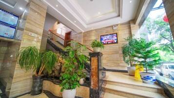 Um saguão de hotel com piso de mármore, escada, plantas em vasos e uma tela de televisão.