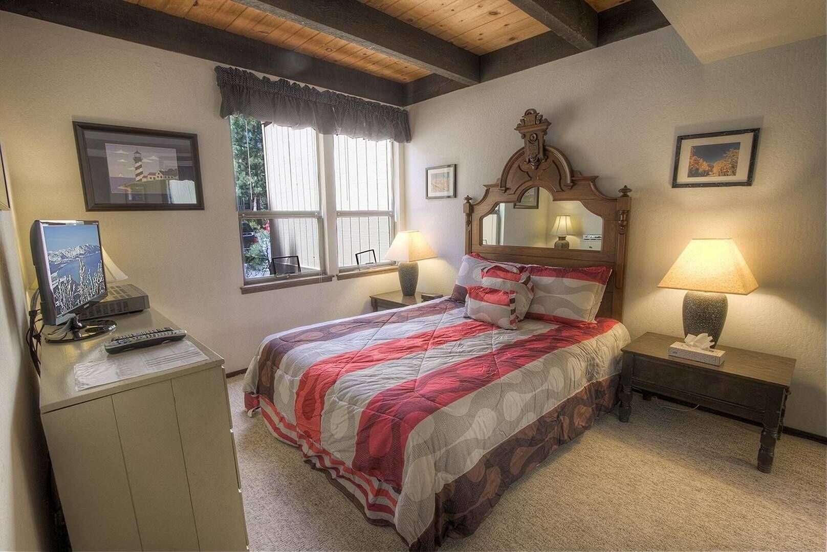 Un dormitorio con cama, un escritorio con computadora, dos lámparas de noche y una ventana con vista a árboles.