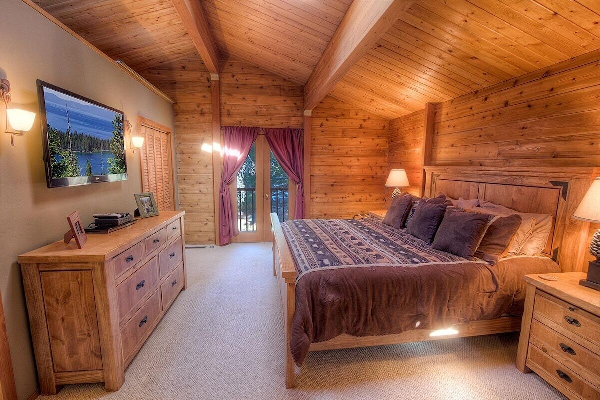 A wooden cabin bedroom with a bed, bedside tables, a dresser, a TV, and a window with curtains.
