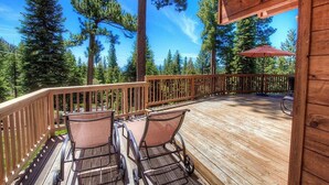 A wooden deck with two chairs and a table, surrounded by tall trees. - The Bass Chalet 3 Bedroom Home by RedAwning (Kings Beach)