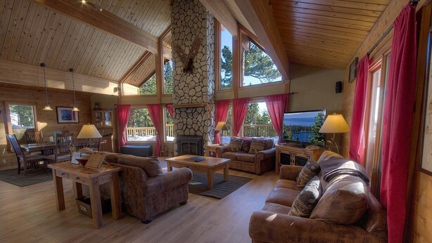 A spacious living room with wooden floors, a stone fireplace, and large windows with red curtains. - The Bass Chalet 3 Bedroom Home by RedAwning (Kings Beach)