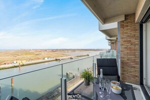 A balcony with a view of a river, a table with glasses and a bowl, and a potted plant. - Lighthouse View-pier facing apartment with garage (Nieuwpoort)