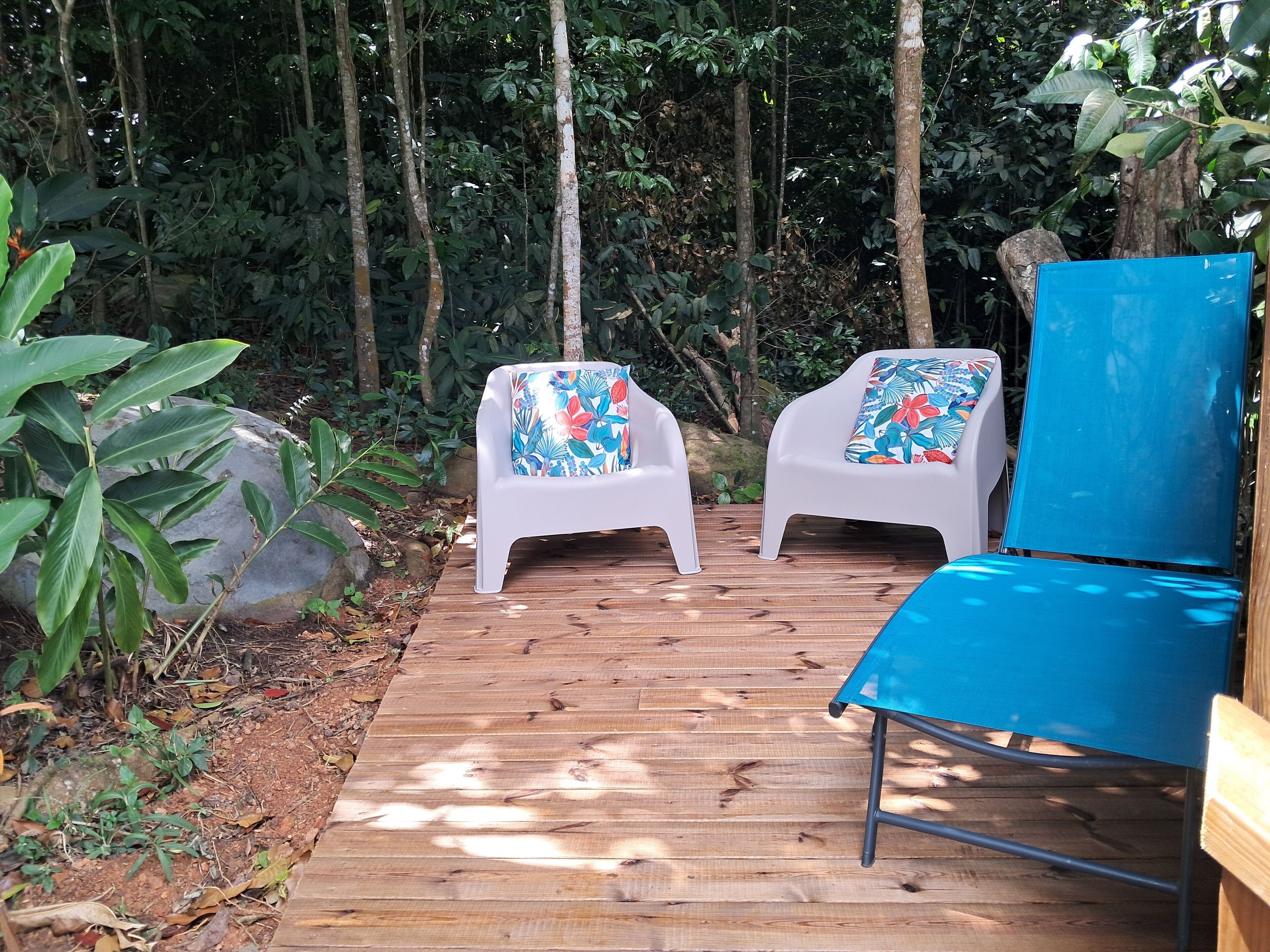 Een houten terras met twee witte plastic stoelen en een blauwe stoel, omgeven door weelderige begroeiing.