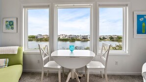 Ein Essbereich mit einem weißen Tisch und Stühlen, großen Fenstern mit Blick auf einen See und Häuser sowie einem grünen Sofa.