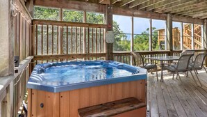 Een houten terras met een jacuzzi, buitenmeubels en uitzicht op de omliggende bomen.