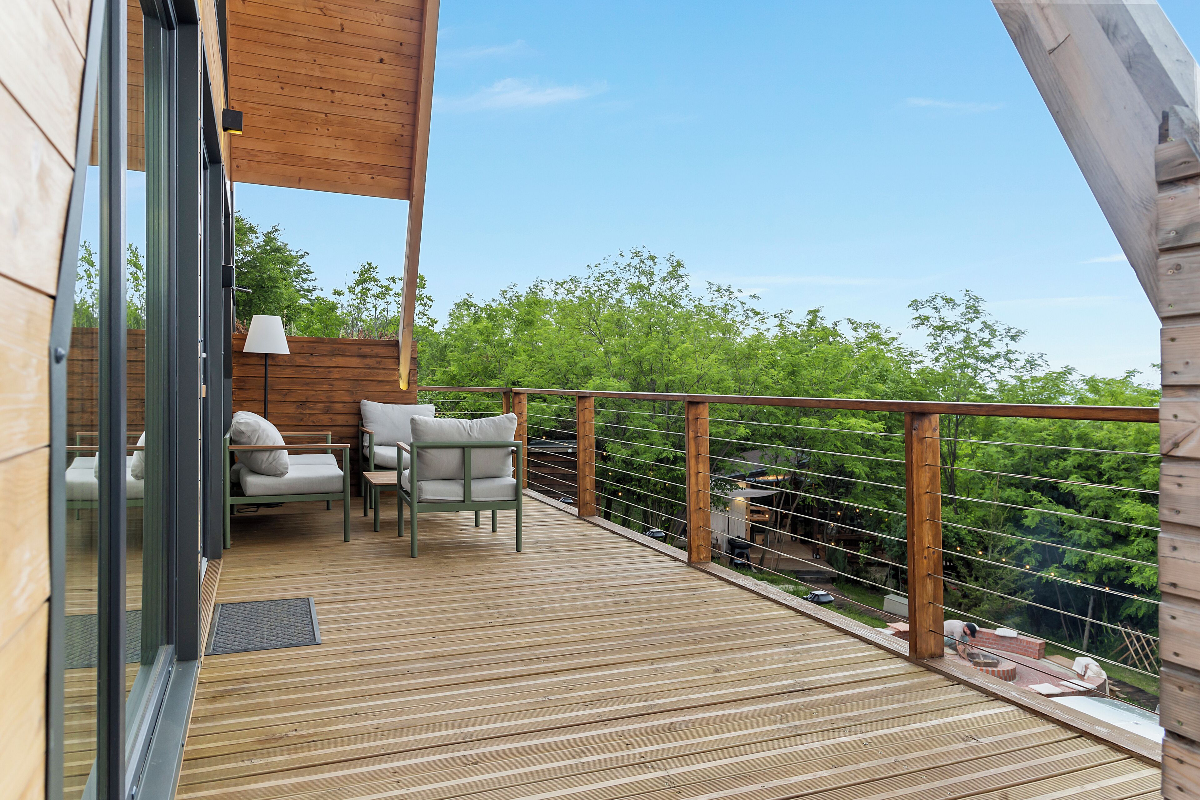 Una terraza de madera con mobiliario exterior, una puerta de cristal y vista a  árboles.