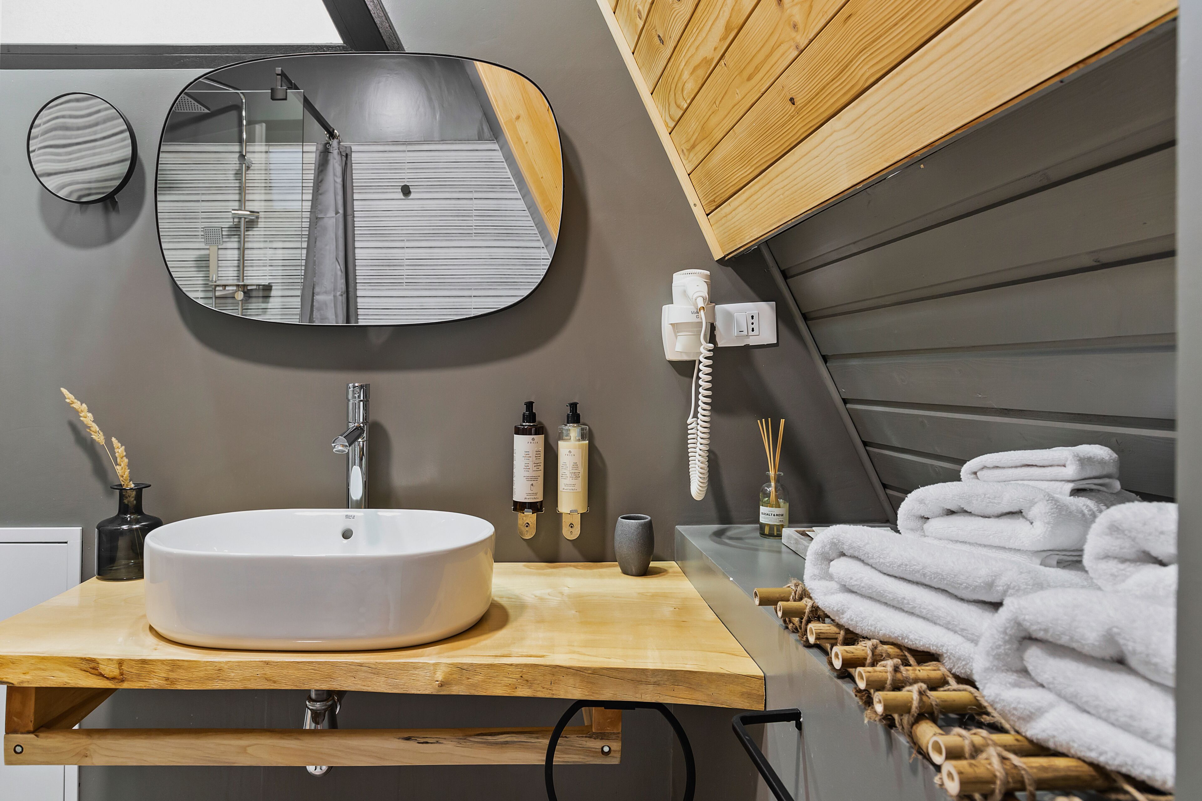 Une salle de bain avec un miroir rond, un lavabo blanc, un meuble-lavabo en bois, un porte-serviettes et un espace douche.