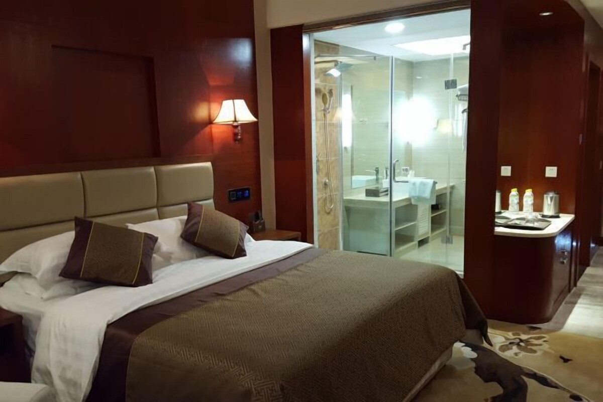 A hotel room with a bed, a nightstand, a lamp, a glass shower enclosure, and a small table with toiletries.