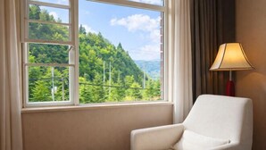 A room with a white armchair, a small round table, and a window offering a view of a forested hillside. - Jiaozishan Resort Hotel (Luquan)