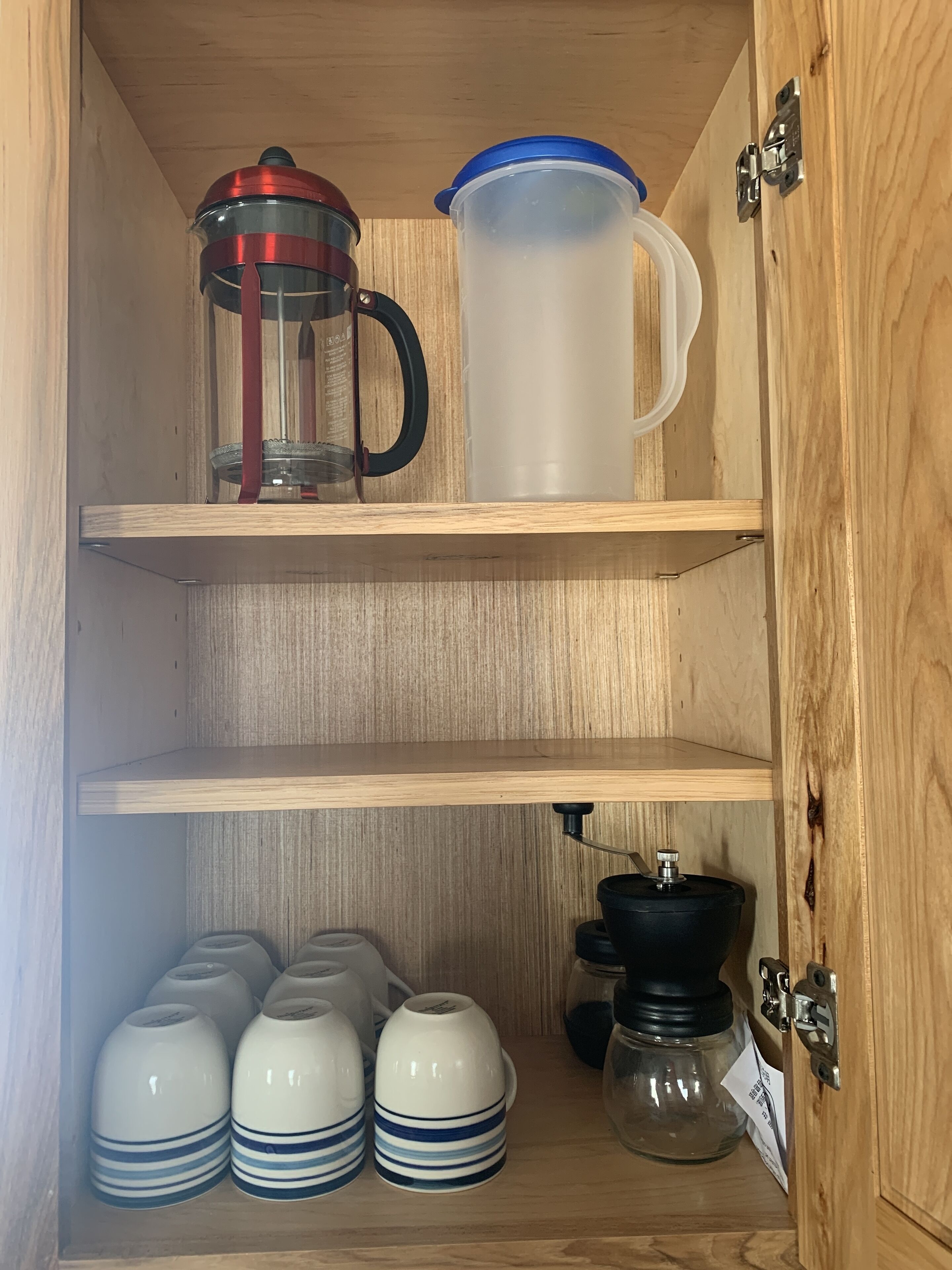 A wooden cabinet with shelves holding a coffee maker, a pitcher, and several white mugs with blue stripes.