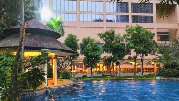 Área de piscina de hotel com um gazebo, palmeiras e um prédio com várias janelas.