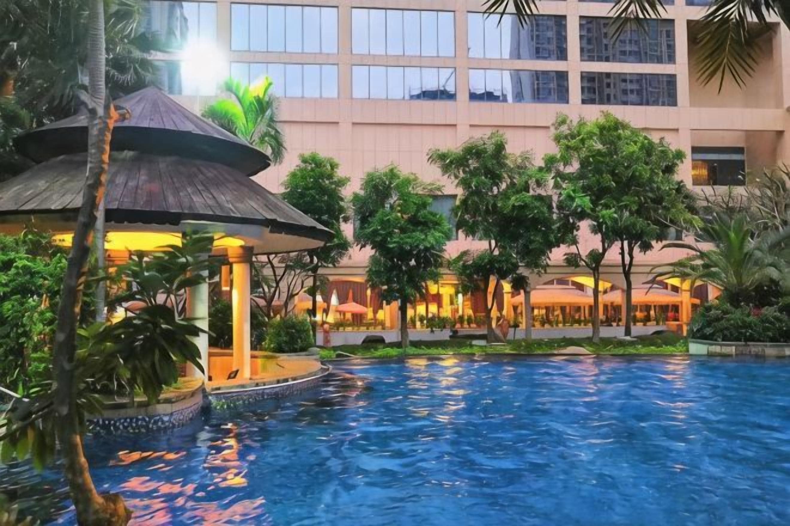 A swimming pool with a gazebo, surrounded by trees and a building with multiple floors.