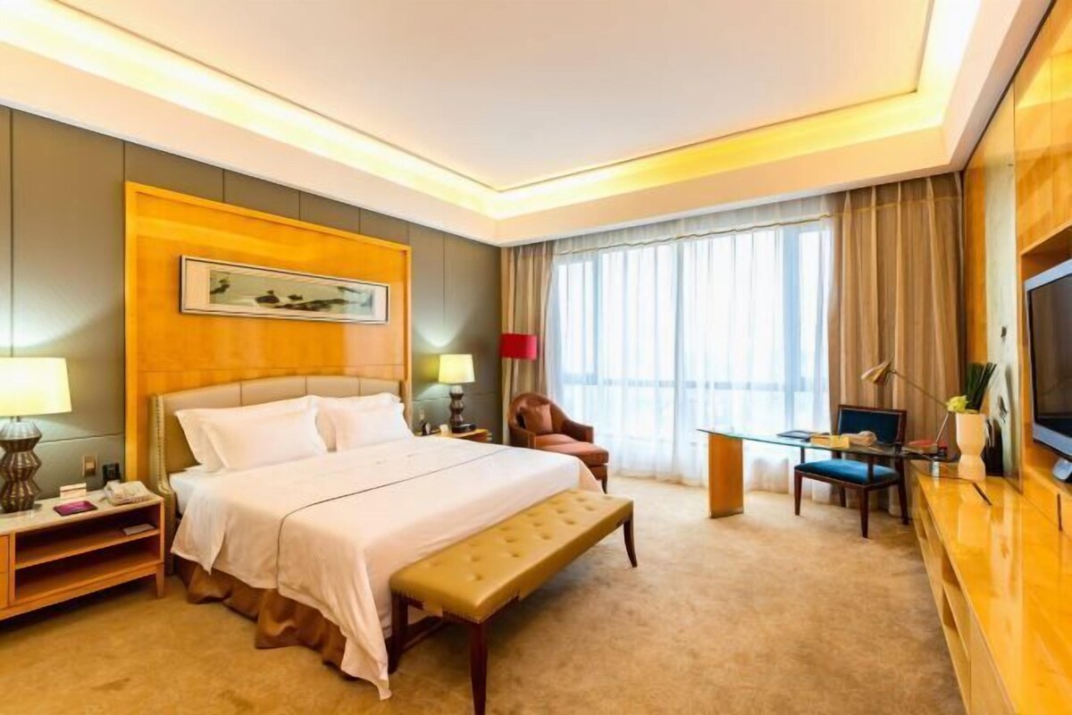 Una habitación de hotel con cama, mesita de noche, lámpara, silla, televisión y ventana con cortinas.