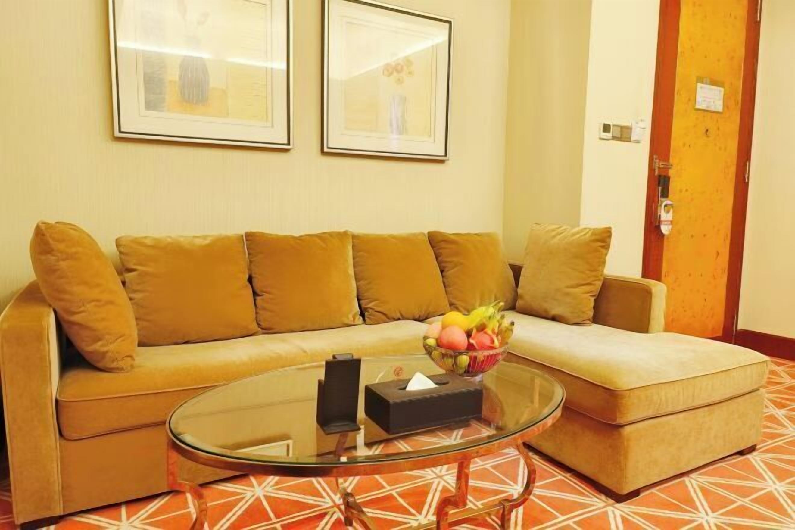 Una sala de estar con un sofá beige, una mesa de centro de vidrio y un frutero. Hay dos cuadros en la pared y una puerta con cerradura.