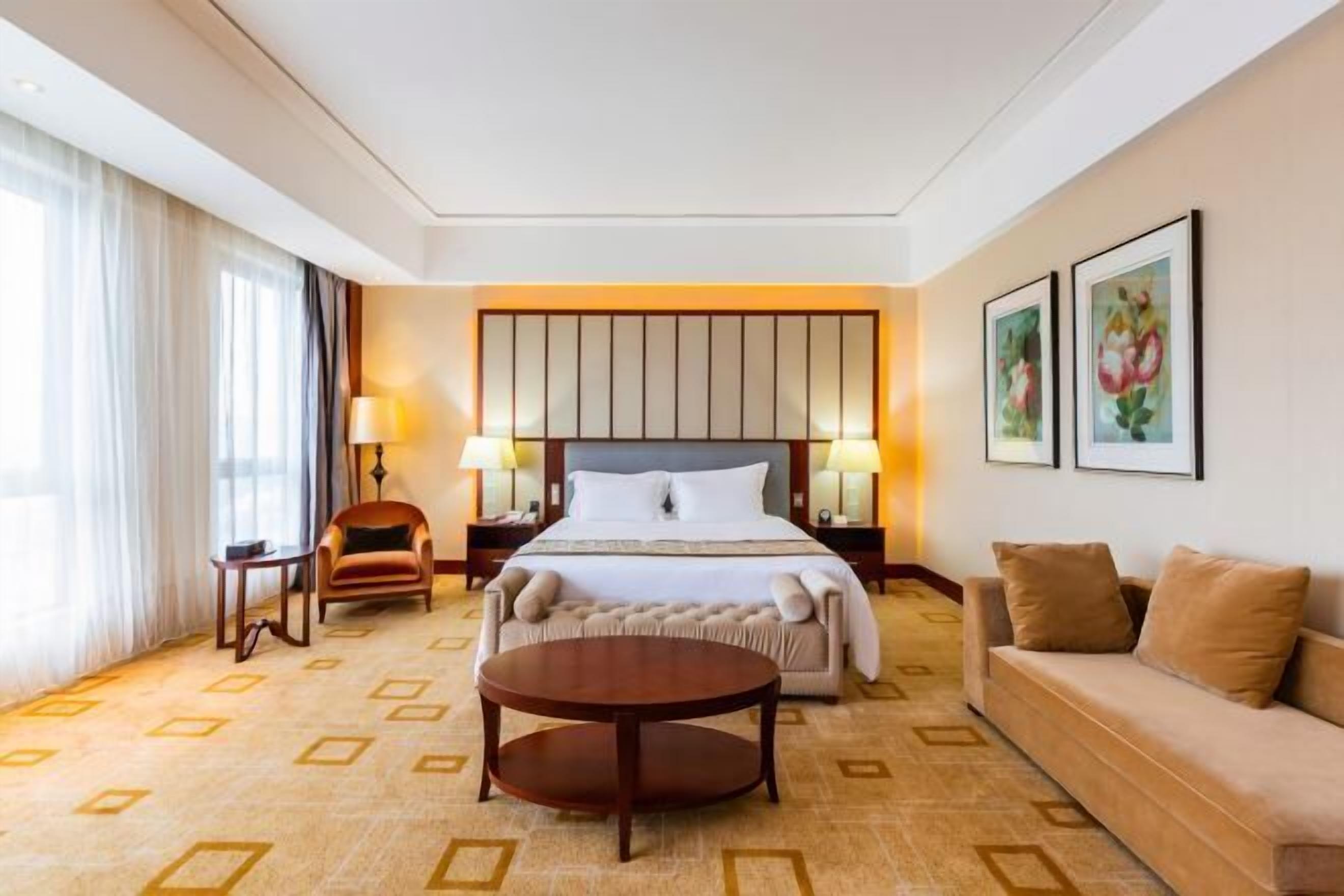 Una habitación de hotel con una cama grande, dos mesitas de noche con lámparas, una mesa de centro redonda, un sofá y una alfombra estampada.