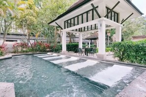 Una piscina con caída de agua, rodeada de abundante vegetación y una zona de estar cubierta.