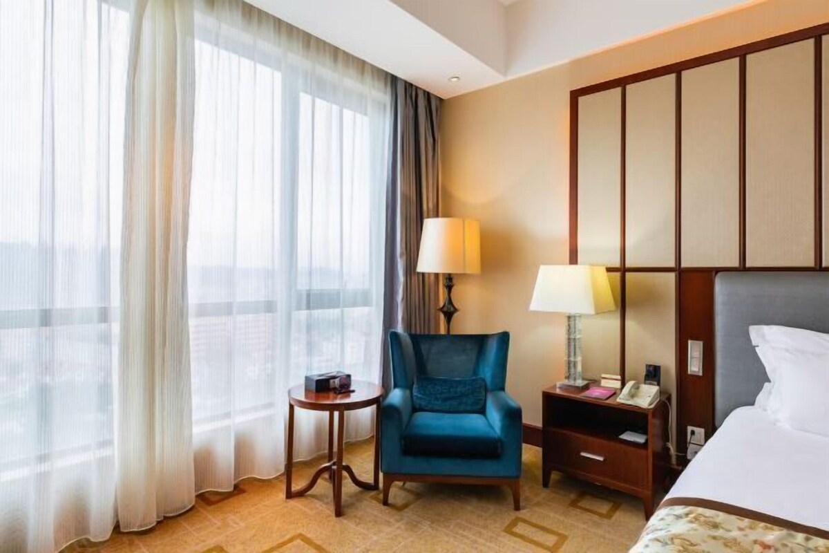 Una habitación de hotel con una cama, un sillón azul, una mesita de noche con una lámpara, una pequeña mesa y un ventanal con cortinas translúcidas.