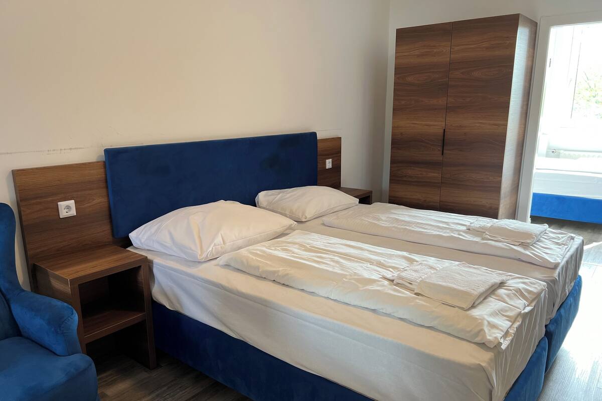 Una cama doble con sábanas blancas, cabecera de madera, mesita de noche y una silla azul.