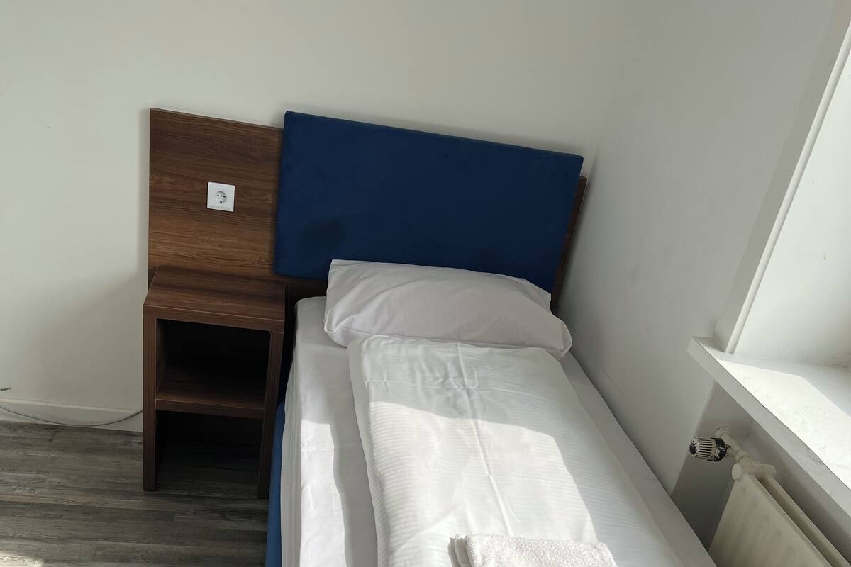 Una cama individual con ropa blanca y un cabecero azul, una mesita de noche de madera con toma de corriente y un radiador en la pared.