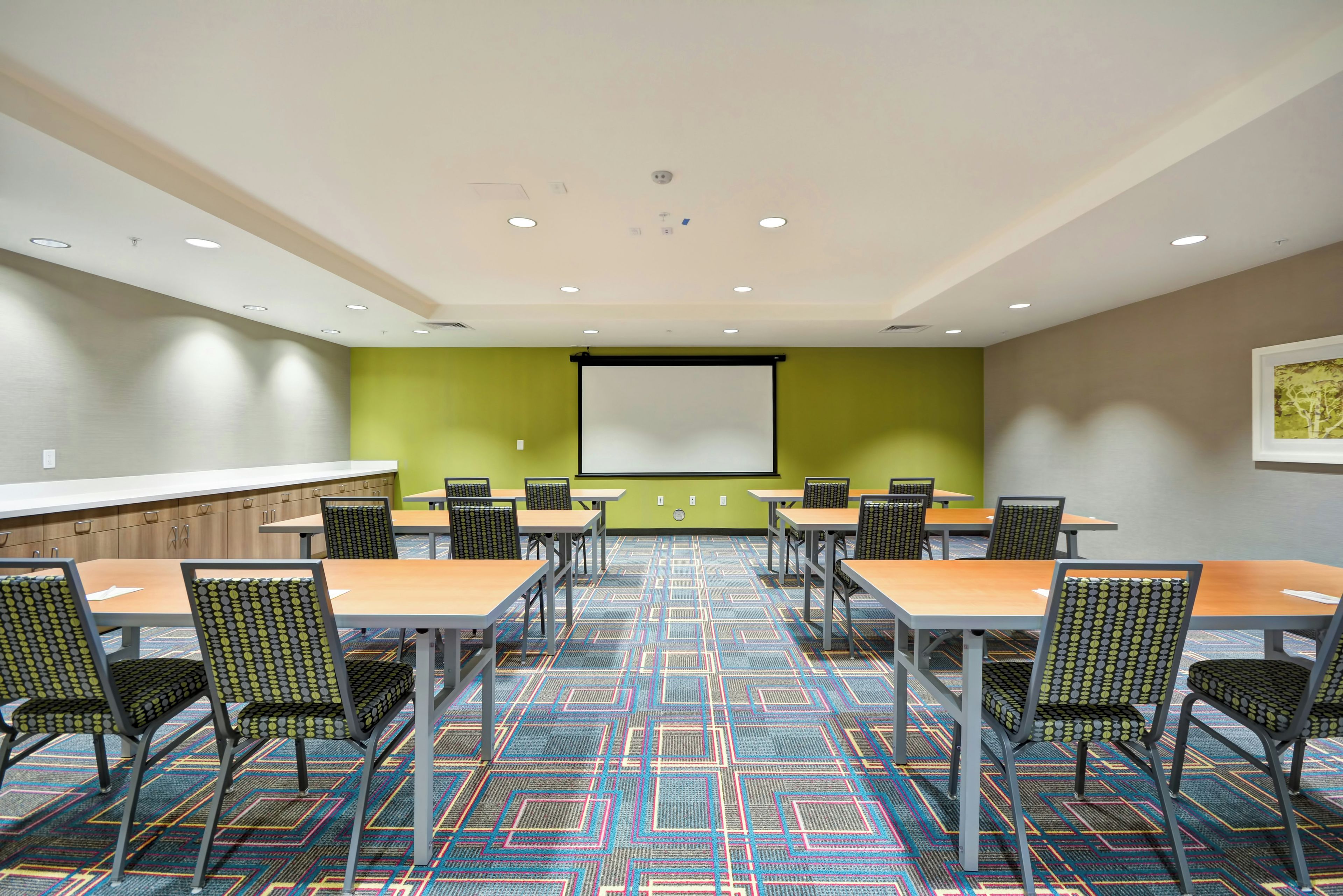 A conference room with a projector screen, multiple tables, chairs, and a patterned carpet.