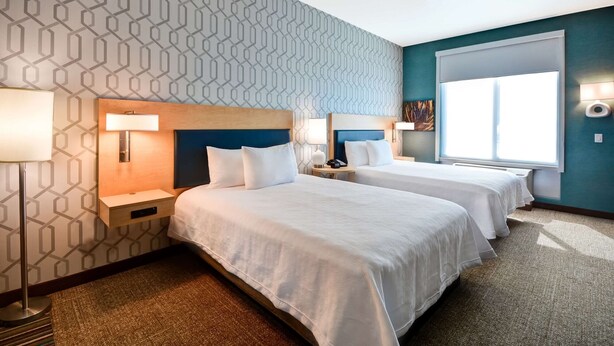 A hotel room with two beds, a patterned wall, a large window, and a nightstand with a lamp. - Home2 Suites By Hilton Livermore (Livermore)