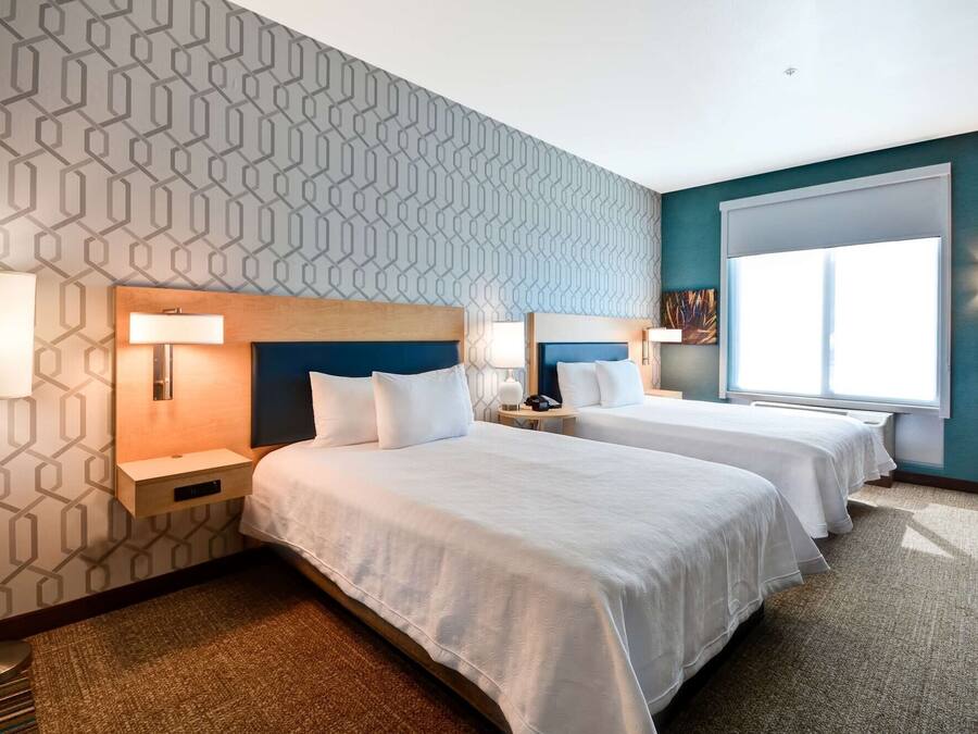 Quarto de hotel com duas camas, parede estampada, uma janela grande e mesa de cabeceira com abajur.