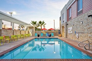 Une piscine bordée d’un muret en pierre, avec des chaises vertes et rouges, et des parasols.