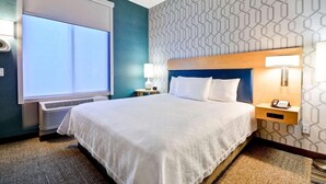 A hotel room with a large bed, a nightstand with a lamp, a wall-mounted phone, and a window with blinds. - Home2 Suites By Hilton Livermore (Livermore)