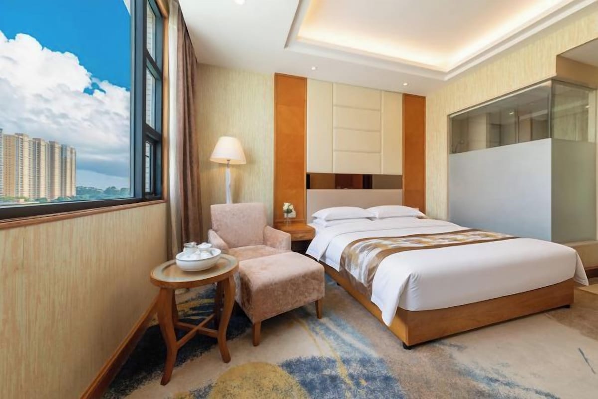 Habitación de hotel con una cama grande, una silla, una mesita y vistas a un paisaje urbano a través de una ventana.