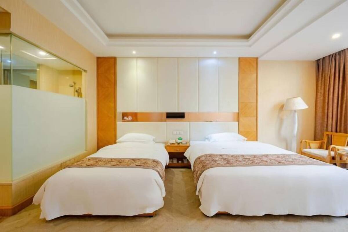 Habitación de hotel con dos camas, un espejo grande, una mesita de noche, una lámpara y vista al baño.