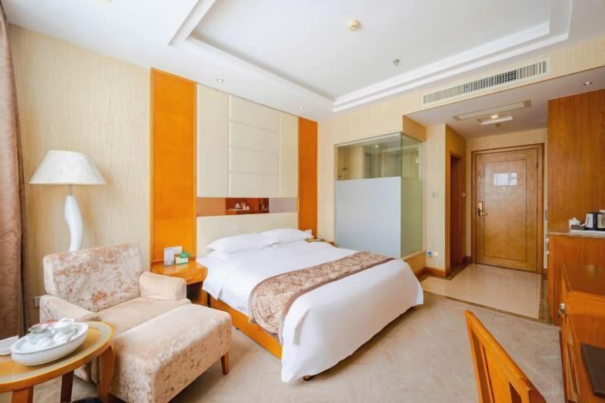 A hotel room with a bed, a chair, a small table, a lamp, and a wardrobe.