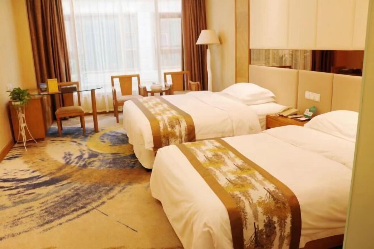 A hotel room with two beds, a desk, chairs, a lamp, and a view of the outdoors.