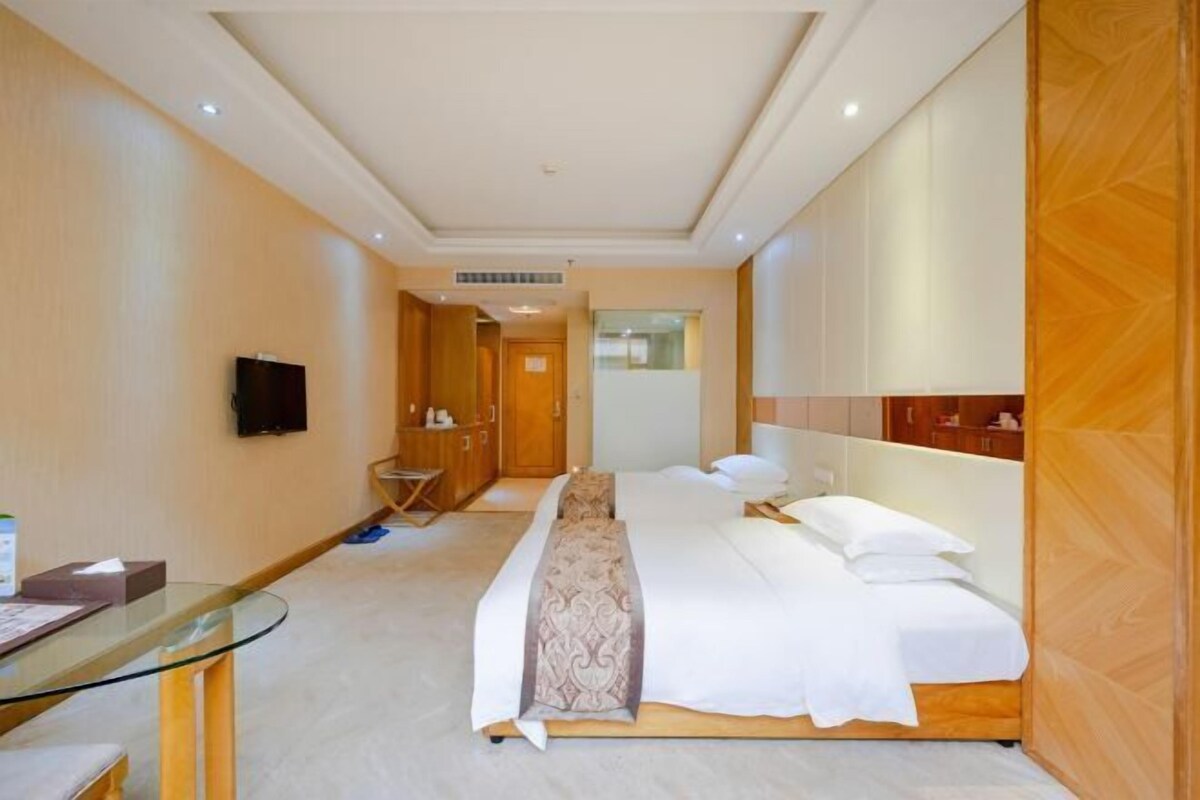 Et hotelværelse med en stor seng, et glastilbord, et fjernsyn ophængt på væggen og en træskab.