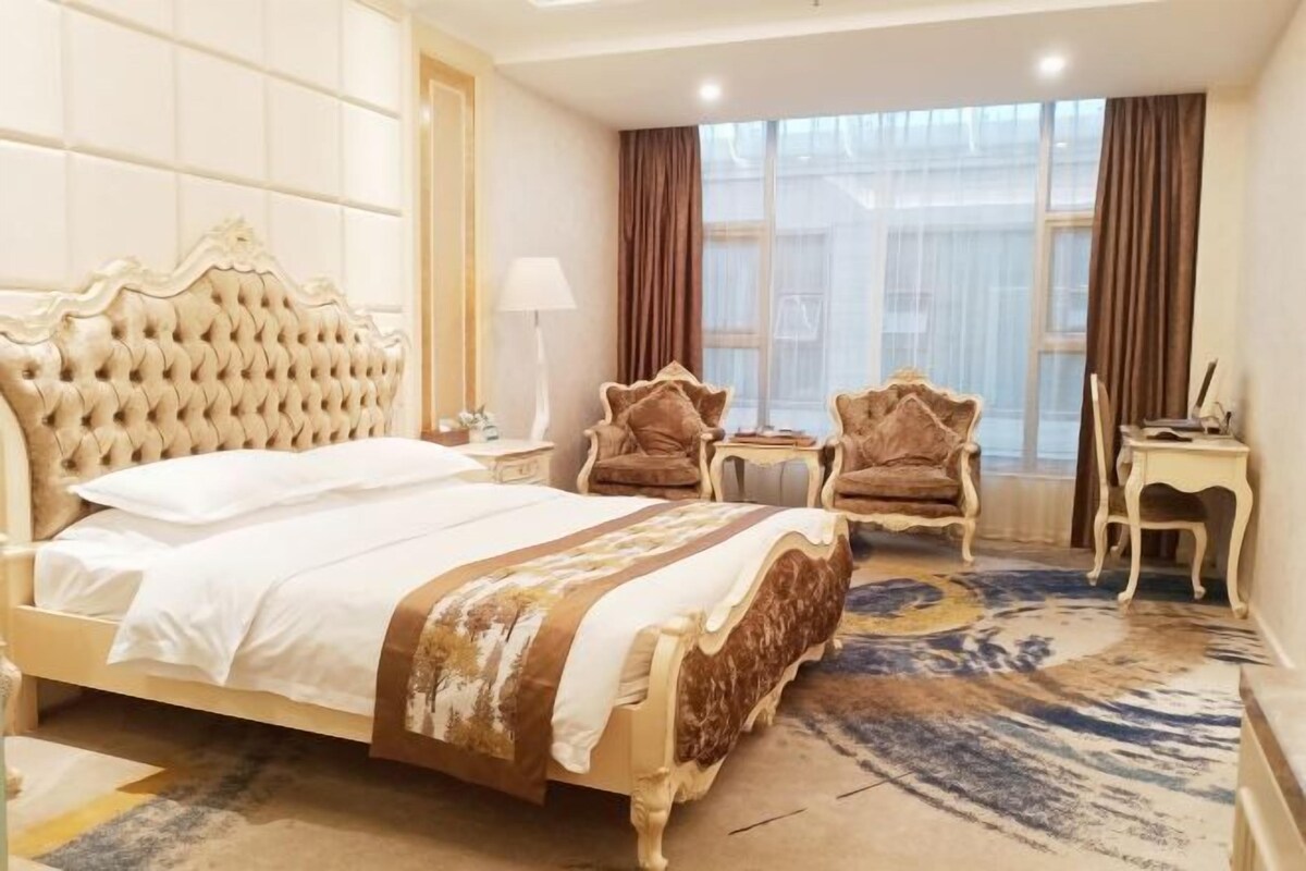 Habitación de hotel con una cama grande, dos sillones, una mesita y un ventanal con cortinas.