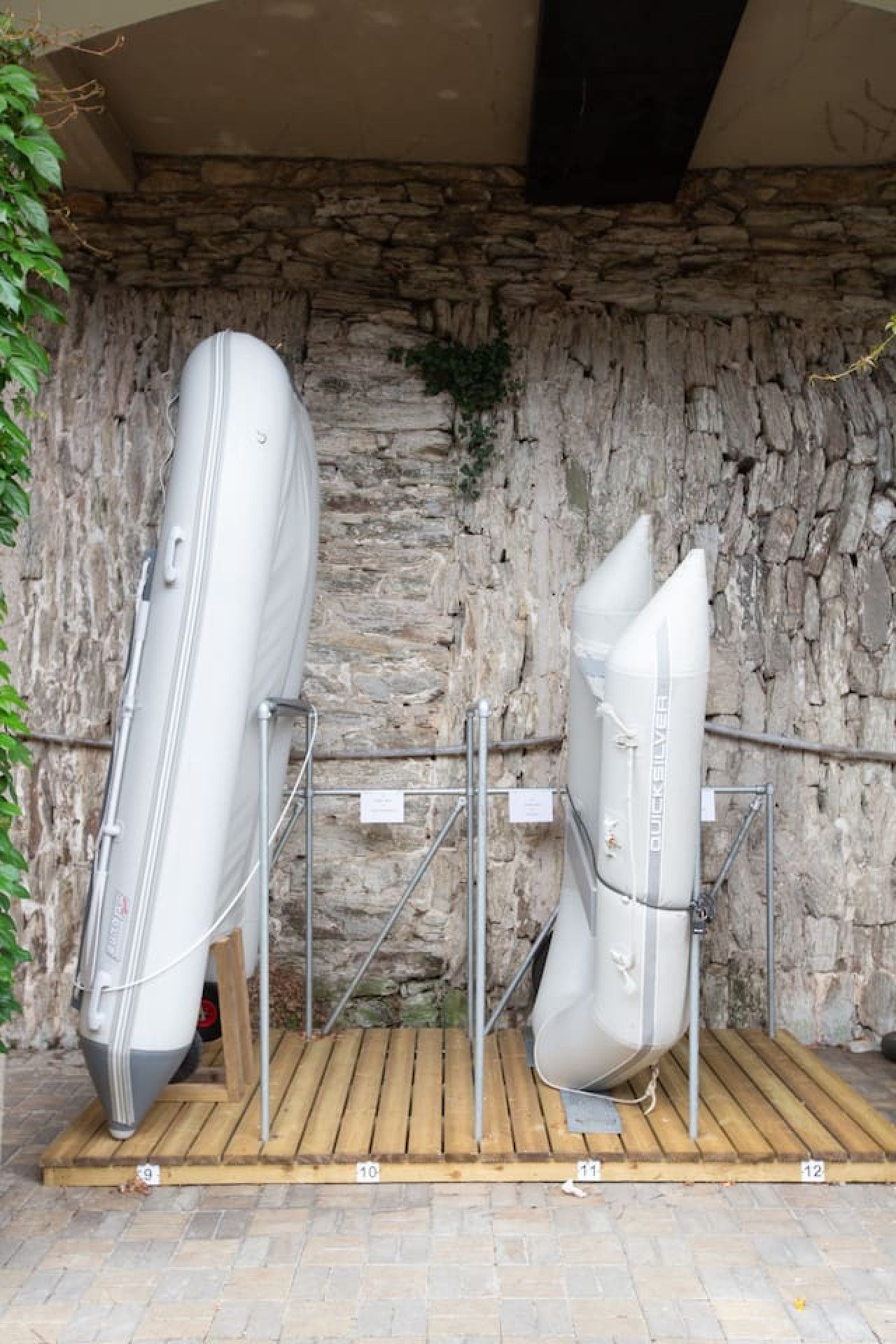 Two white kayaks secured on a wooden platform with metal supports, against a stone wall backdrop.