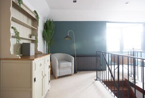 Et værelse med et hvidt skrivebord, en grå lænestol, en gulvlampe og udsigt til en balkon.