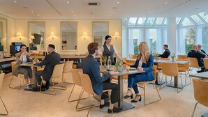 A modern dining area with multiple tables and chairs, a waiter attending to guests, and large windows providing natural light. - Self Check-In Hotel Am Brinkerplatz (Essen)