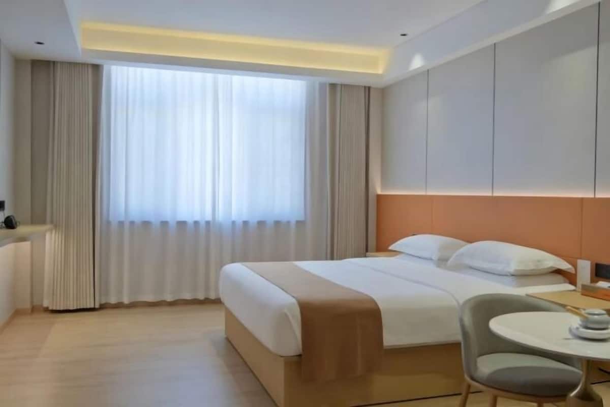 Habitación de hotel con cama, una mesita, una silla y un gran ventanal con cortinas.